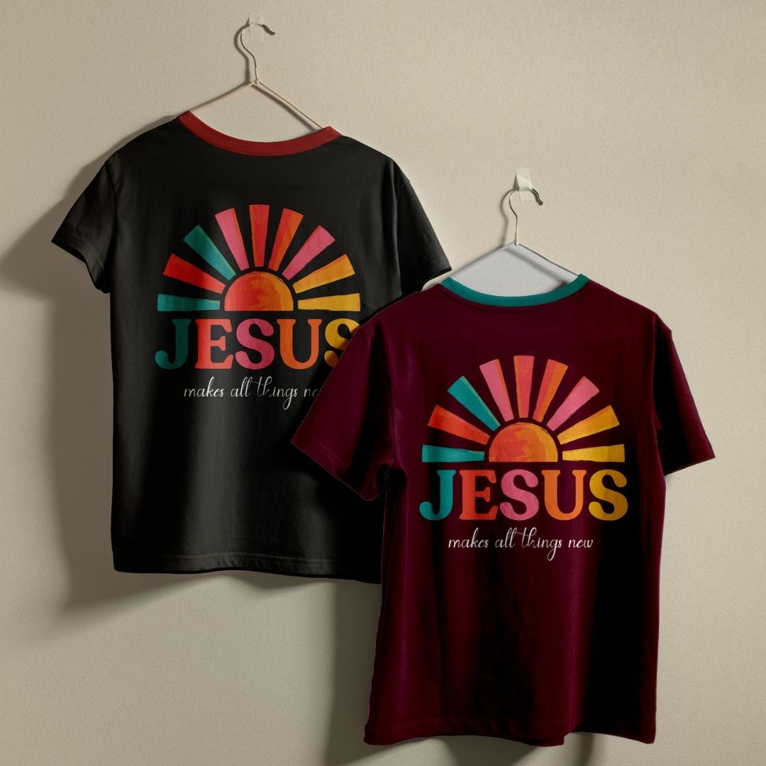 Two short-sleeve graphic T-shirts hanging on hangers against a plain wall. The left shirt is black with a red collar and a multicolor sunburst graphic on the back reading 'JESUS makes all things new.