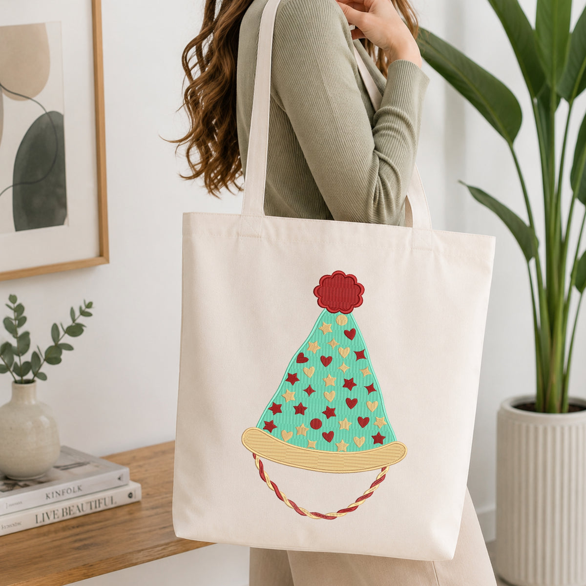 A woman wearing a sage green ribbed sweater carries a cream canvas tote bag with a colorful embroidered party hat design. The background is a bright, modern room with a wooden table, books, a framed print, and a potted plant.