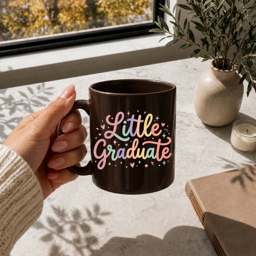 A hand holds a dark brown ceramic mug with a colorful 'Little Graduate' text design, set on a windowsill with a book, candle, and potted plant nearby.