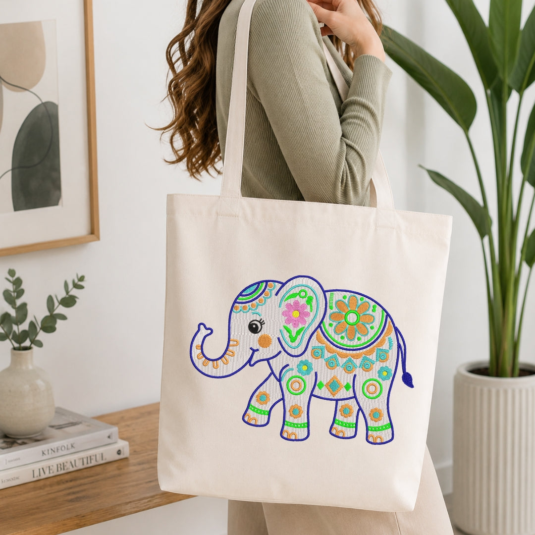 A woman wearing a sage green ribbed sweater carries a cream canvas tote bag with a colorful, patterned elephant design printed on the front, set in a bright, modern room with a wooden table and potted plants.