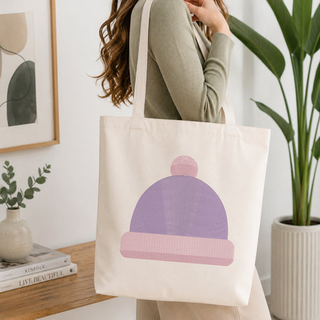 A woman wearing a sage green ribbed sweater carries a cream canvas tote bag featuring a stylized purple and pink knit beanie graphic. The scene is set in a bright, modern room with a wooden table, books, a small plant, and a large green potted plant in the background.