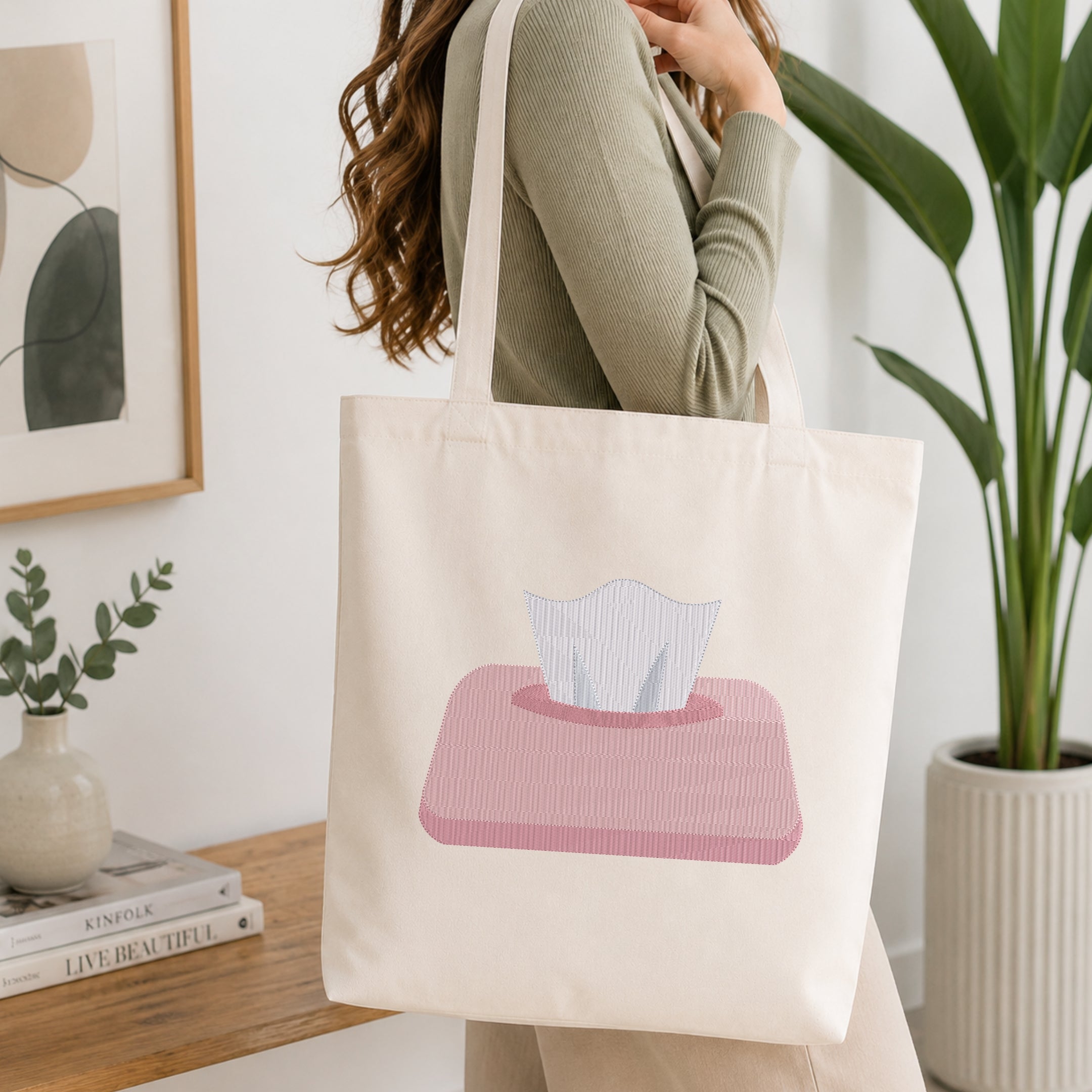 A person holding a natural canvas tote bag printed with an embroidered tissue box design, set against a bright interior room background with green plants.