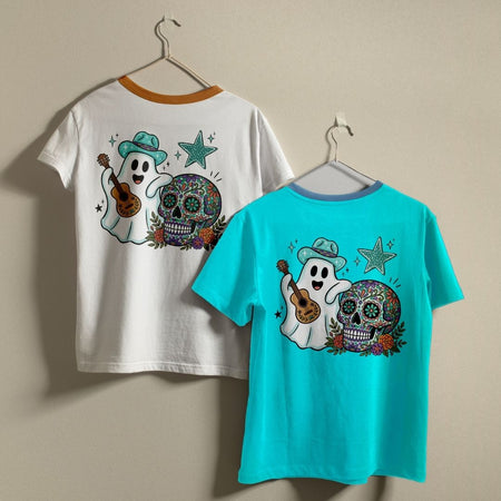 Two children's short-sleeve t-shirts hanging on hangers against a plain beige wall. One shirt is white with a brown collar trim and features a colorful back graphic of a ghost playing a guitar next to a decorated sugar skull.