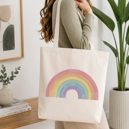 A woman wearing a sage green ribbed long-sleeve top carries a cream canvas tote bag with a colorful rainbow design printed on the front, set in a bright, modern room with a wooden shelf, framed art, and a potted plant.