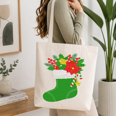A woman wearing a ribbed sage green long-sleeve top carries a cream canvas tote bag decorated with a festive Christmas stocking graphic. The graphic features a green stocking filled with red poinsettias, holly leaves, green pine branches, a yellow ornament, and candy canes.