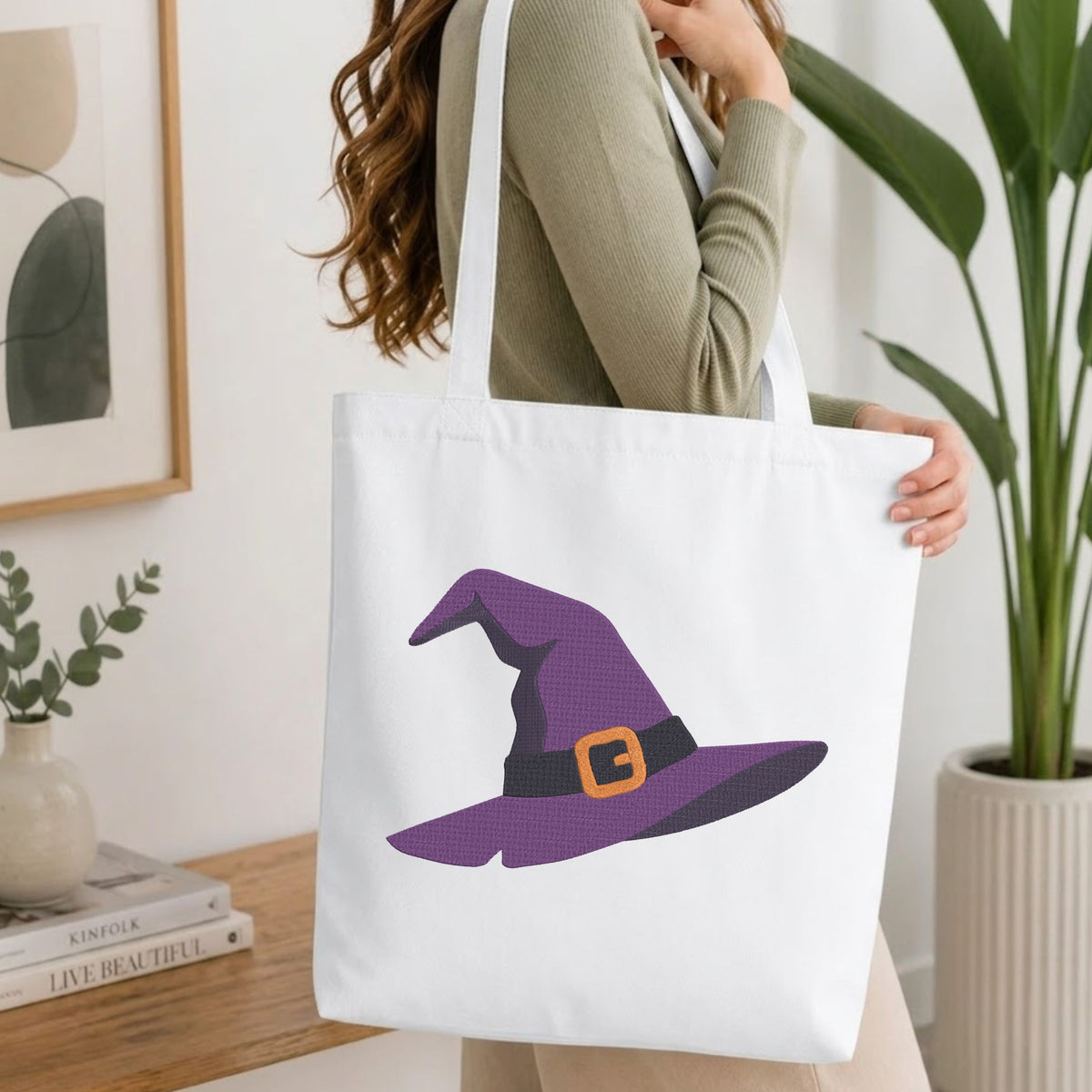A woman wearing a sage green ribbed sweater carries a white canvas tote bag featuring a purple witch hat graphic with a black band and orange buckle, set against a bright, modern indoor background with a wooden table and potted plants.