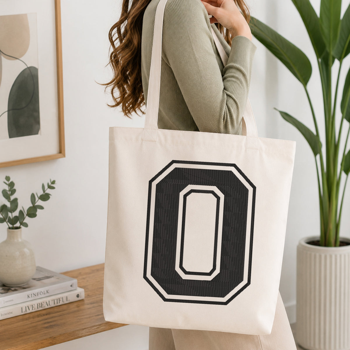 A woman wearing a sage green ribbed long-sleeve top carries a large off-white canvas tote bag with a bold black collegiate-style letter 'O' graphic on the front, set in a bright, modern room with a wooden console table and a potted plant.