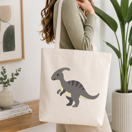 A woman wearing a sage green ribbed sweater carries a cream canvas tote bag with a gray and yellow dinosaur graphic. The scene is set in a bright, modern room with a wooden table, books, a small plant, and a large green potted plant in the background.