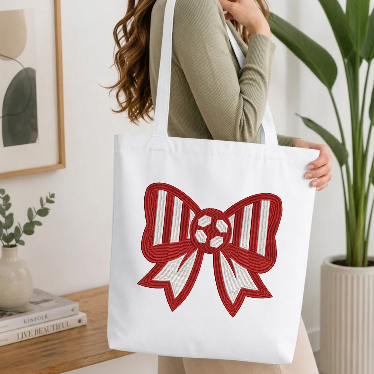 Woman carrying a white natural canvas tote bag featuring an embroidered red and white soccer bow design in a bright, modern interior setting.