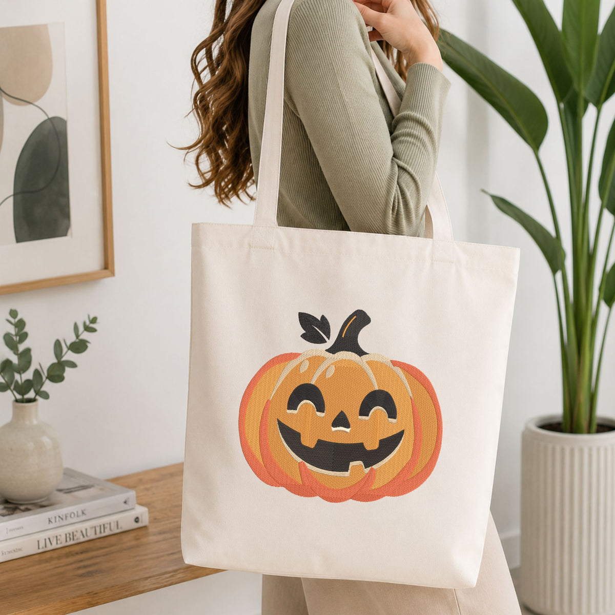 Woman carrying a white natural canvas tote bag featuring an embroidered jack-o'-lantern pumpkin design in a bright, modern interior setting.