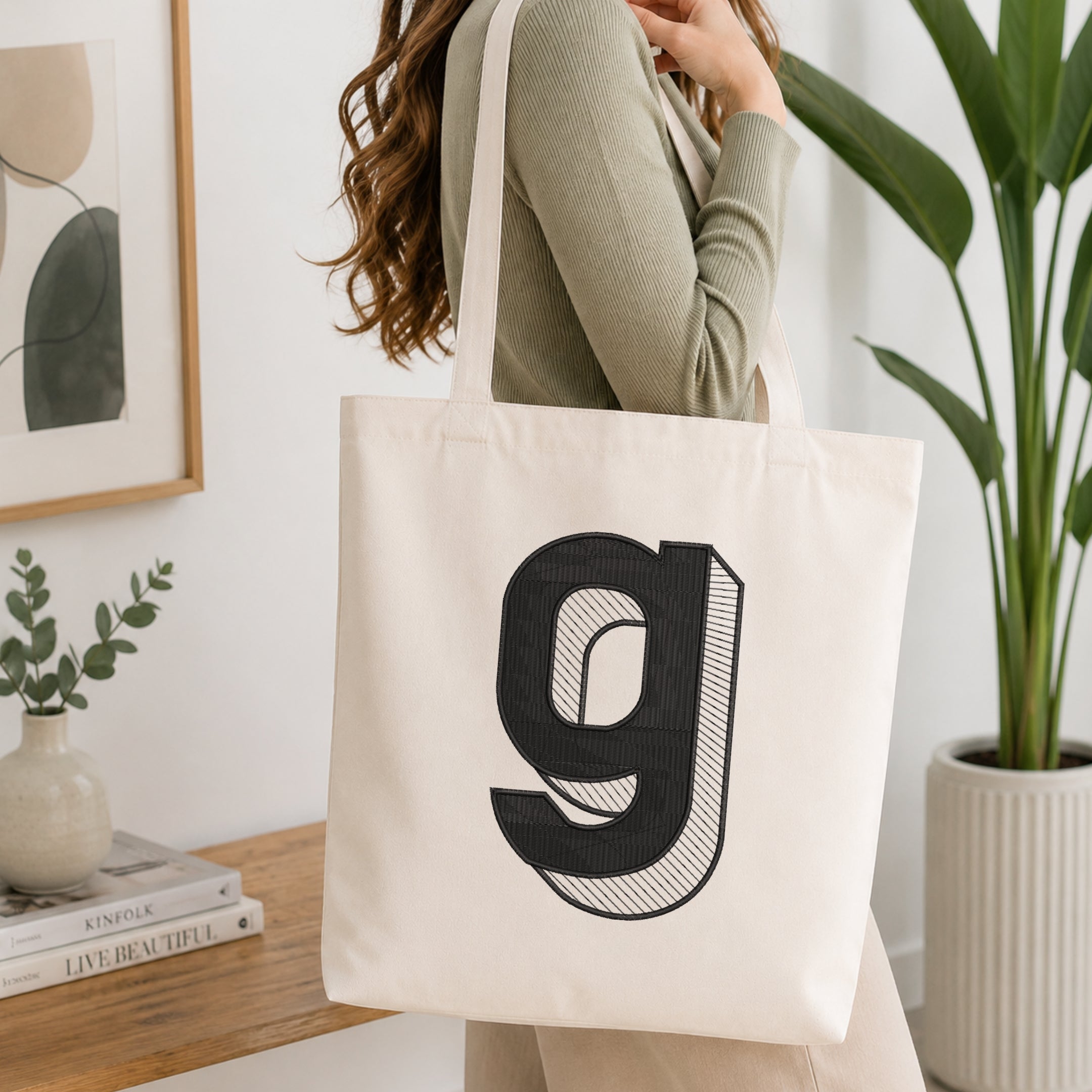 A person holding a natural canvas tote bag printed with an embroidered 3D block lowercase letter "g" design, set against a bright interior room background with green plants.
