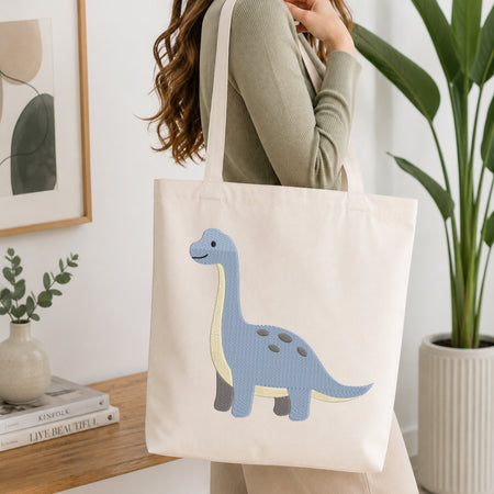 A woman wearing a sage green ribbed sweater carries a cream canvas tote bag with an embroidered blue long-neck dinosaur on the front, set in a bright, modern room with a wooden table, books, and a potted plant.