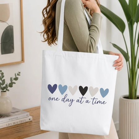 A woman wearing a sage green ribbed long-sleeve top carries a white canvas tote bag with six heart graphics in navy, light blue, black, beige, and light blue, above the text 'one day at a time' in navy script.