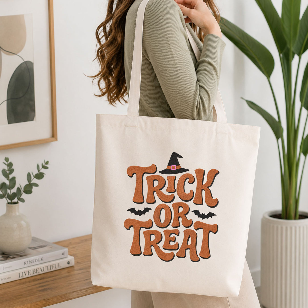 Woman carrying a white natural canvas tote bag featuring a "Trick or Treat" witch hat machine embroidery design in a bright, modern interior setting   