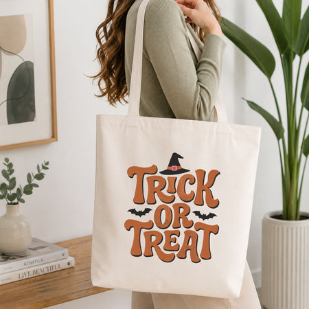 Woman carrying a white natural canvas tote bag featuring a "Trick or Treat" witch hat machine embroidery design in a bright, modern interior setting   