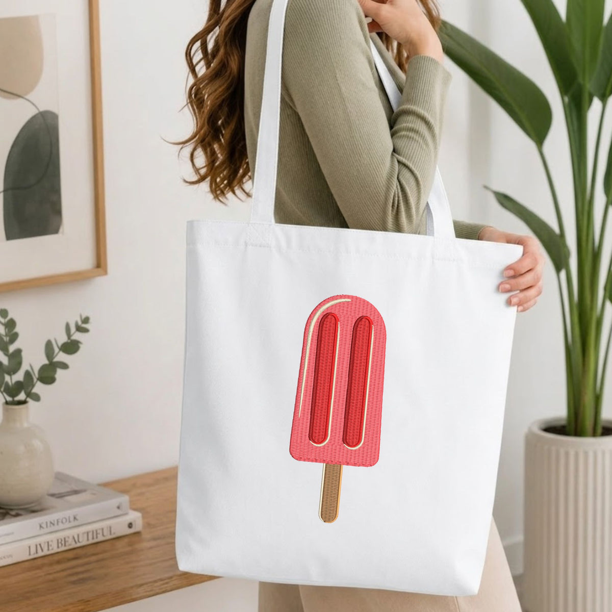  Woman carrying a white natural canvas tote bag featuring an embroidered pink ice cream popsicle design in a bright, modern interior setting.   