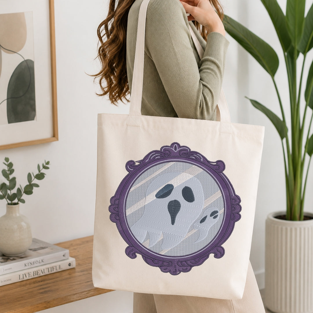 A person wearing a sage green ribbed long-sleeve top carries a cream canvas tote bag with a purple-framed ghost illustration on the front, set in a bright, modern room with a wooden table, books, and a potted plant.