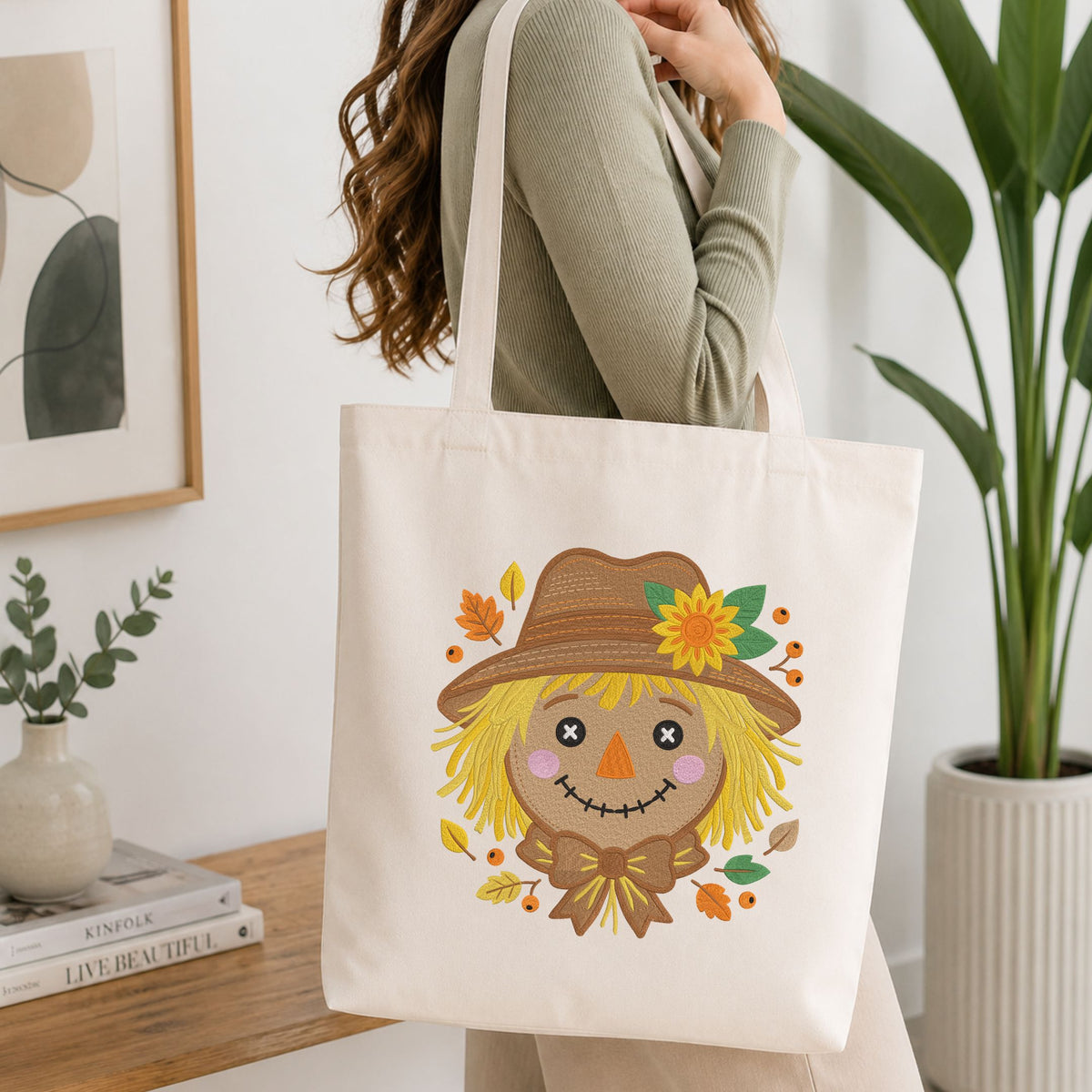 Woman carrying a white natural canvas tote bag featuring a cute autumn scarecrow face machine embroidery design in a bright, modern interior setting