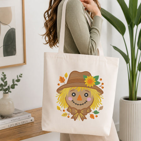 Woman carrying a white natural canvas tote bag featuring a cute autumn scarecrow face machine embroidery design in a bright, modern interior setting