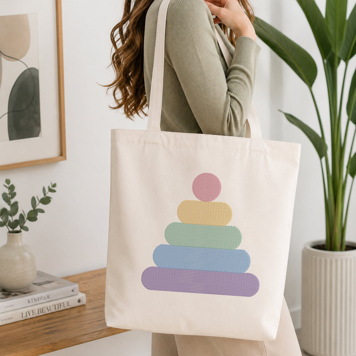 A woman wearing a sage green ribbed sweater carries a cream canvas tote bag with a pastel rainbow stack-of-cups graphic. The setting is a bright, modern room with a wooden table, a potted green plant, and a framed abstract print.