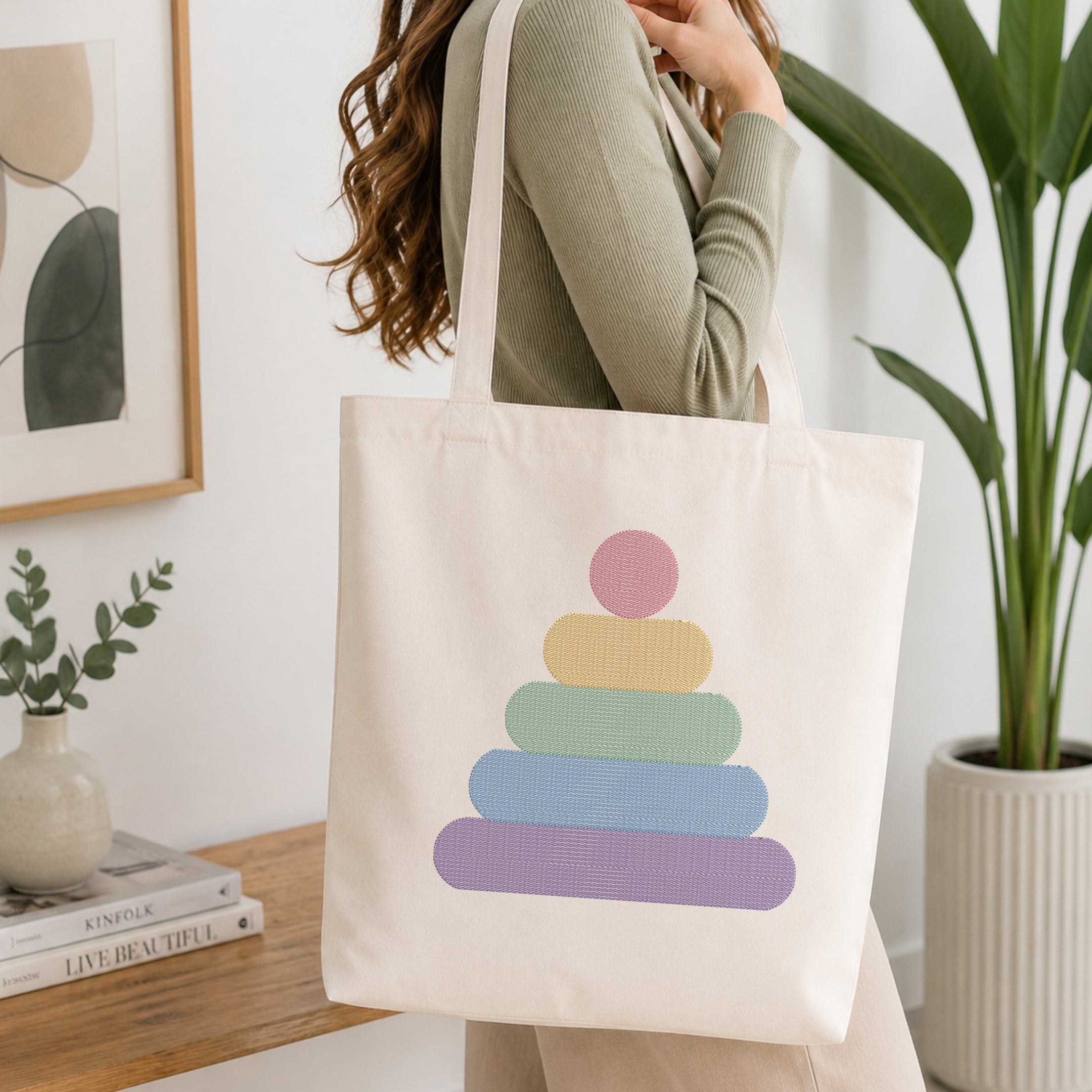 A woman wearing a sage green ribbed sweater carries a cream canvas tote bag with a pastel rainbow stack-of-cups graphic. The setting is a bright, modern room with a wooden table, a potted green plant, and a framed abstract print.
