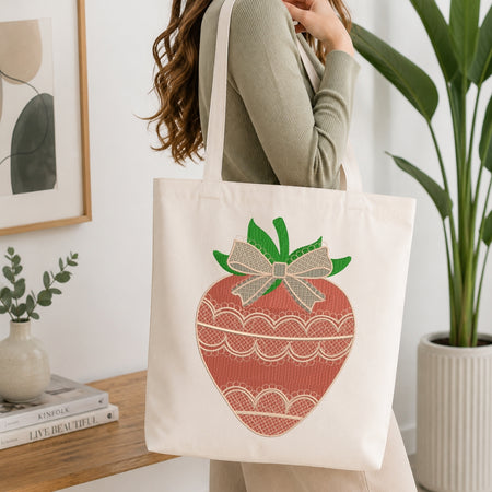 A woman wearing a sage green ribbed sweater carries a cream canvas tote bag featuring a large embroidered strawberry design in terracotta red, green, and beige.