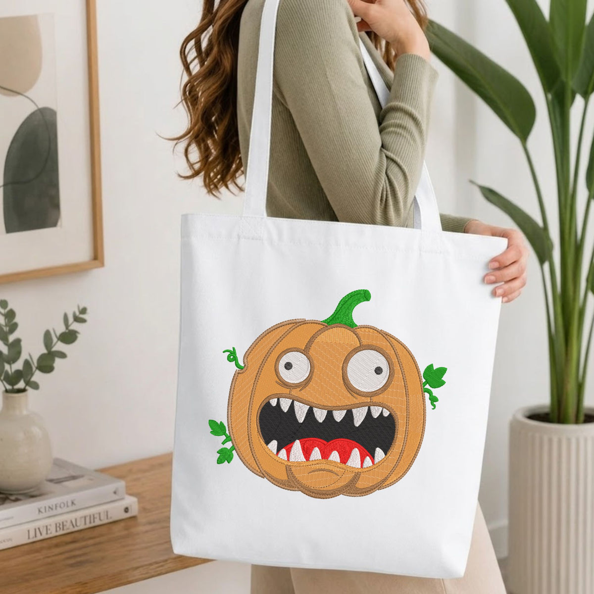 A woman wearing a sage green ribbed long-sleeve top carries a white canvas tote bag with a cartoon pumpkin graphic. The pumpkin has a wide-open mouth showing sharp white teeth, a green stem, and small green vines.