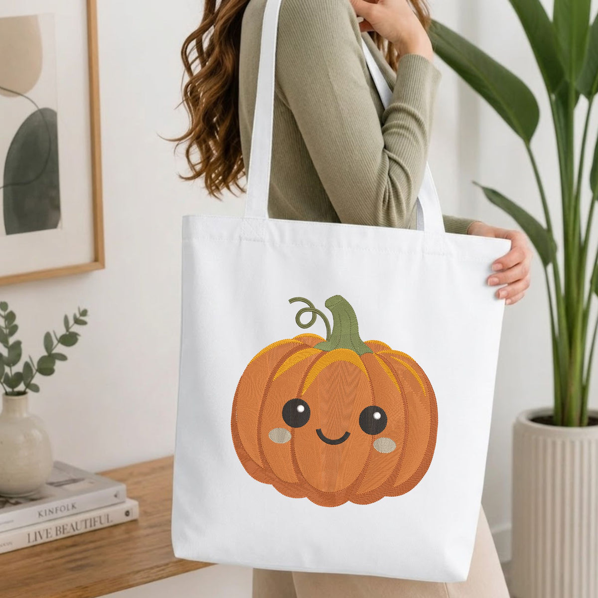 Woman carrying a white natural canvas tote bag featuring an embroidered cute smiling pumpkin design in a bright, modern interior setting.
