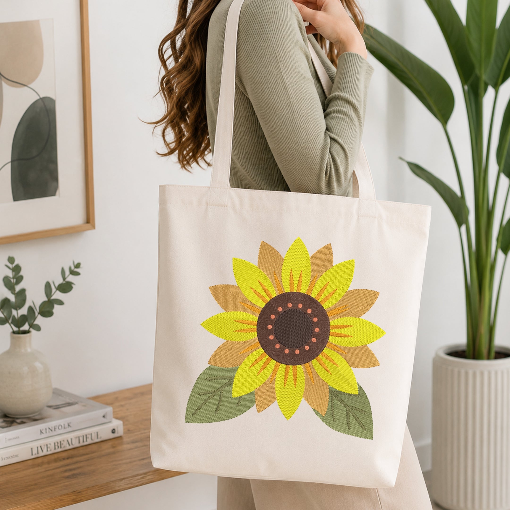 A woman wearing a sage green ribbed long-sleeve top carries a cream canvas tote bag with a large embroidered sunflower design. The background is a bright, modern room with a wooden shelf, framed art, and a potted green plant.
