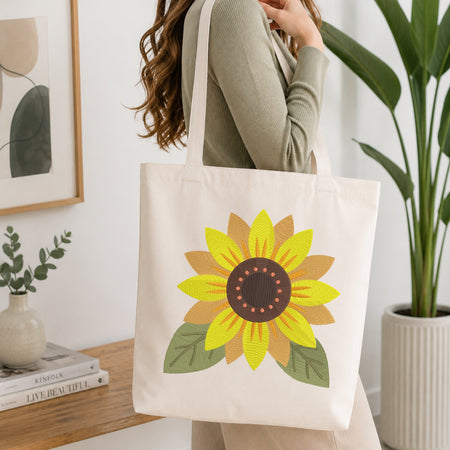 A woman wearing a sage green ribbed long-sleeve top carries a cream canvas tote bag with a large embroidered sunflower design. The background is a bright, modern room with a wooden shelf, framed art, and a potted green plant.