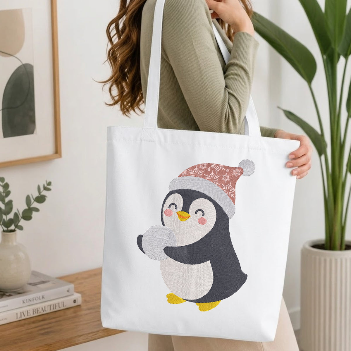 A woman holds a white canvas tote bag featuring a cute cartoon penguin wearing a pink and white patterned Santa hat and holding a gray ball. The scene is set in a bright, modern room with a wooden table, books, and a potted plant in the background.