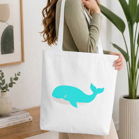  Woman carrying a white natural canvas tote bag featuring an embroidered turquoise blue whale design in a bright, modern interior setting.   