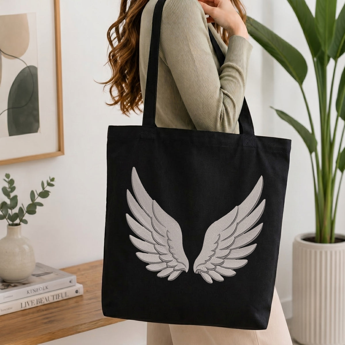 A woman wearing a beige ribbed sweater carries a black canvas tote bag with a large white embroidered angel wings design on the front, set in a bright, modern room with a wooden table, framed art, and a potted plant.