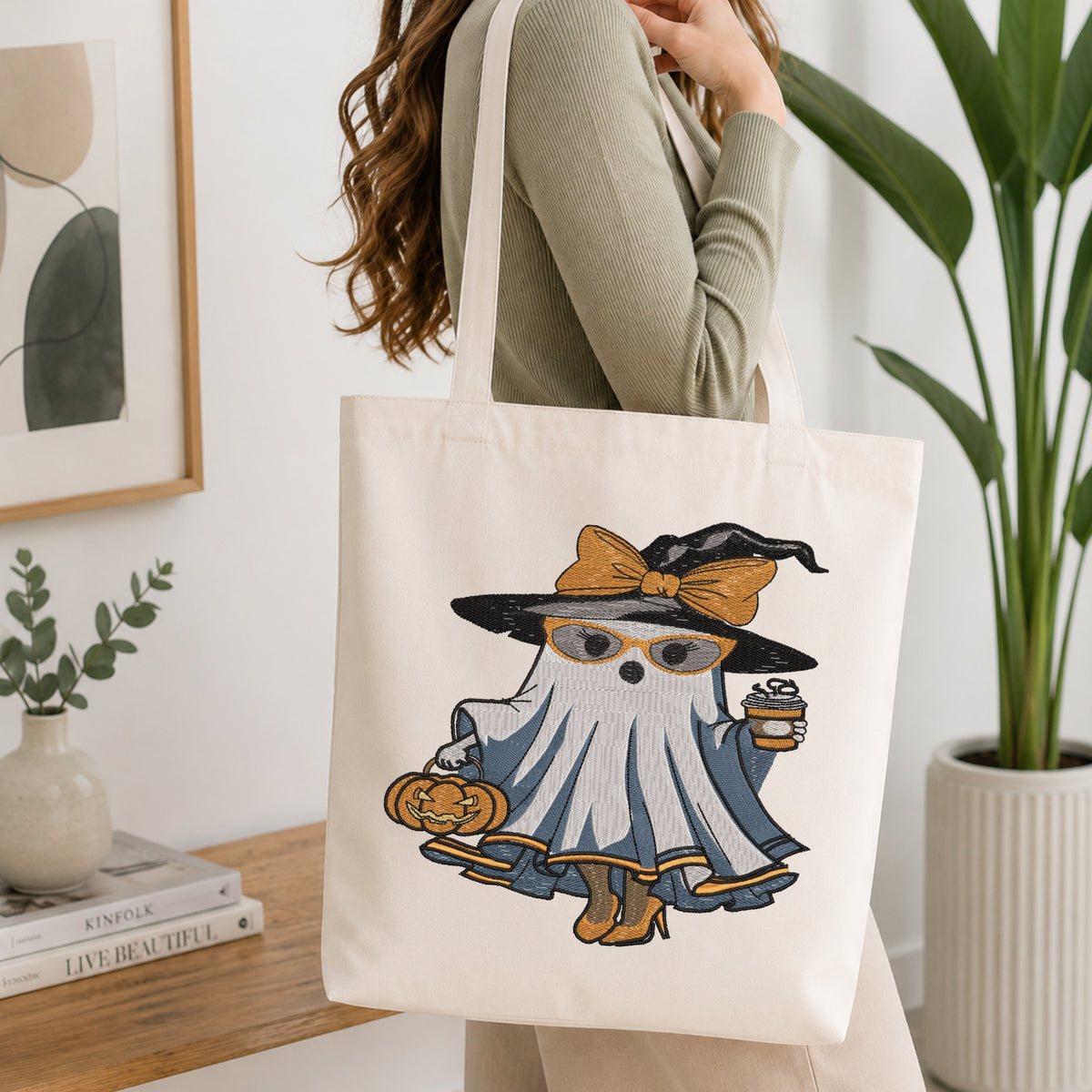 Person holding a canvas tote bag decorated with an embroidery design of a fashionable witch ghost carrying coffee and a pumpkin bucket.