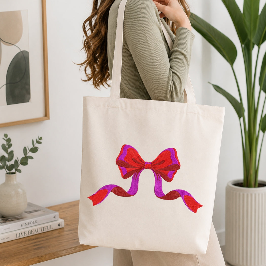 A person wearing a sage green ribbed sweater carries a cream canvas tote bag with a large red and purple bow graphic printed on the front, set in a bright, modern room with a wooden table and potted plants.