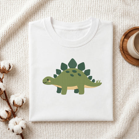 A folded white crewneck t-shirt featuring a cute green dinosaur graphic, styled on a textured cream knit blanket with cotton bolls and a wooden cup in the background.