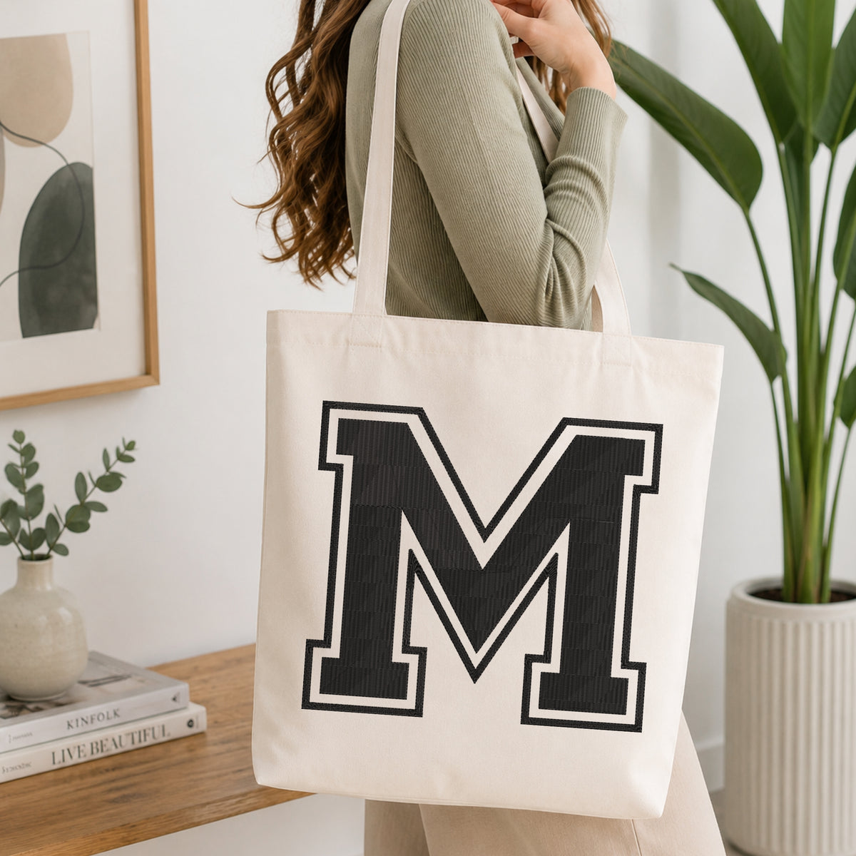 A woman wearing a sage green ribbed sweater carries a cream canvas tote bag with a large black block letter 'M' outlined in white on the front, set in a bright, modern room with a wooden console table and a potted plant.
