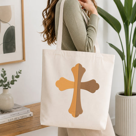 A woman wearing a sage green ribbed long-sleeve top carries a cream canvas tote bag with a large textured cross design in shades of tan and brown on the front. The scene is set in a bright, modern room with a wooden console table, a potted green plant, and framed wall art.
