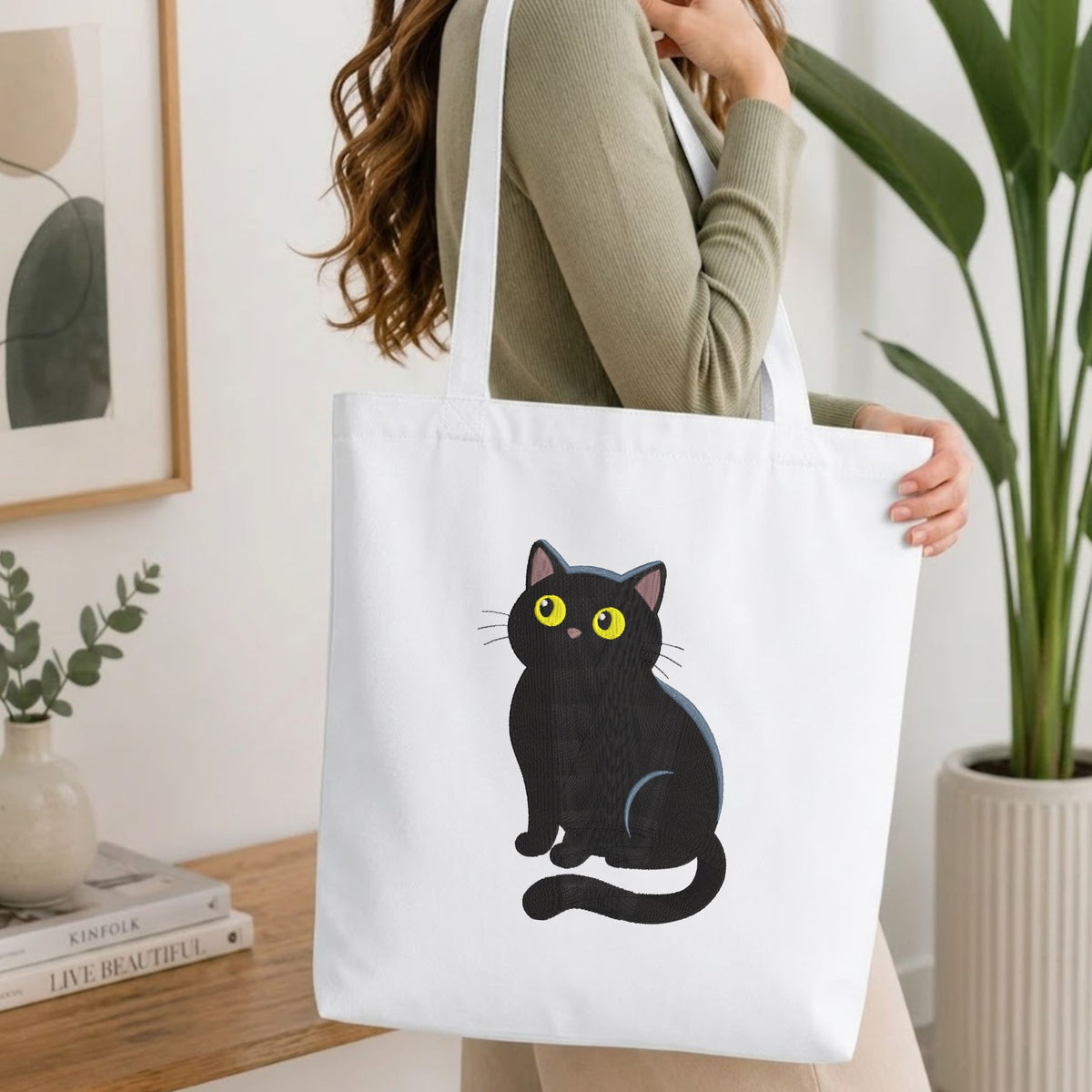 A woman wearing a sage green ribbed sweater carries a white canvas tote bag with a cartoon illustration of a black cat with yellow eyes. The setting is a bright, modern room with a wooden table, books, a framed print, and a potted plant.