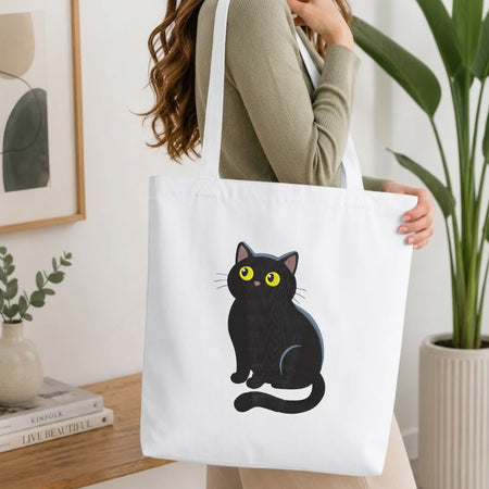 A woman wearing a sage green ribbed sweater carries a white canvas tote bag with a cartoon illustration of a black cat with yellow eyes. The setting is a bright, modern room with a wooden table, books, a framed print, and a potted plant.