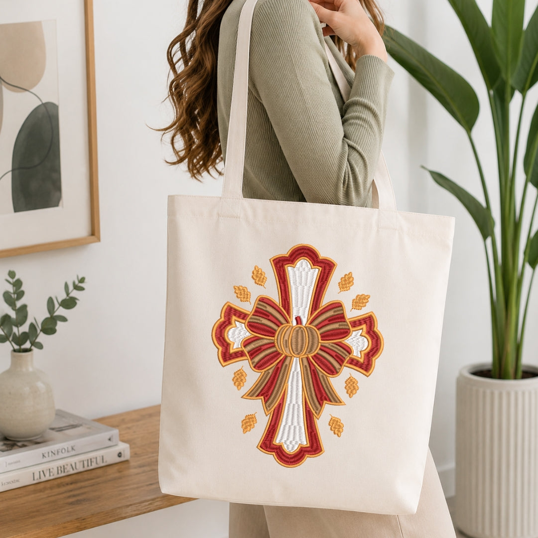 A person wearing a sage green ribbed long-sleeve top carries a cream canvas tote bag with a large embroidered cross design in red, gold, and white, set against a bright, modern interior background with a potted plant and books.