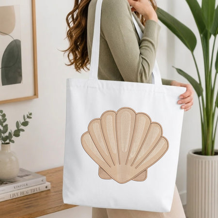 A woman wearing a sage green ribbed long-sleeve top carries a white canvas tote bag featuring a large beige seashell graphic on the front. The setting is a bright, modern room with a wooden table, books, a framed print, and a potted green plant.