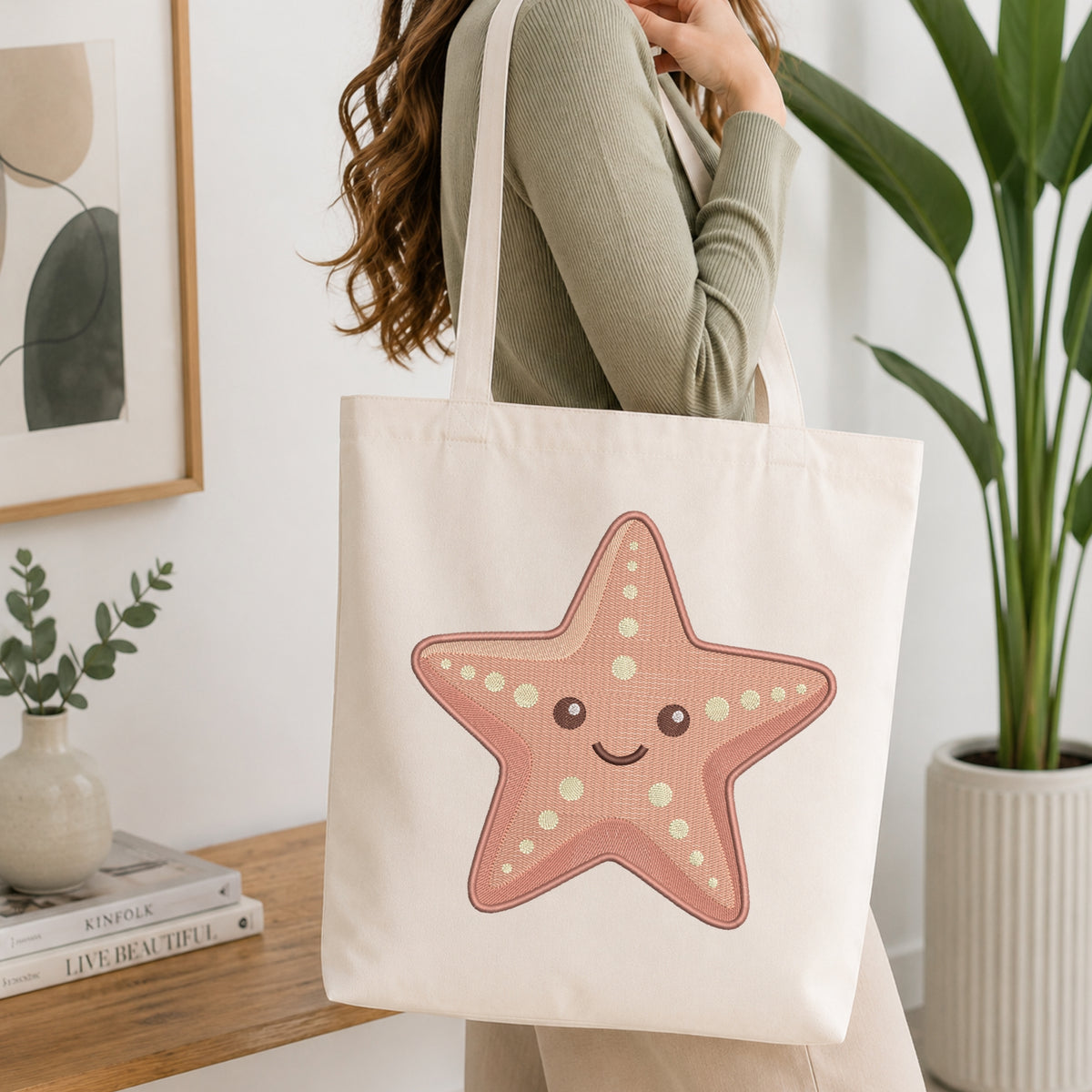 A woman wearing a sage green ribbed sweater carries a cream canvas tote bag with a cute pink starfish graphic printed on the front, set in a bright, modern room with a wooden table and potted plants.
