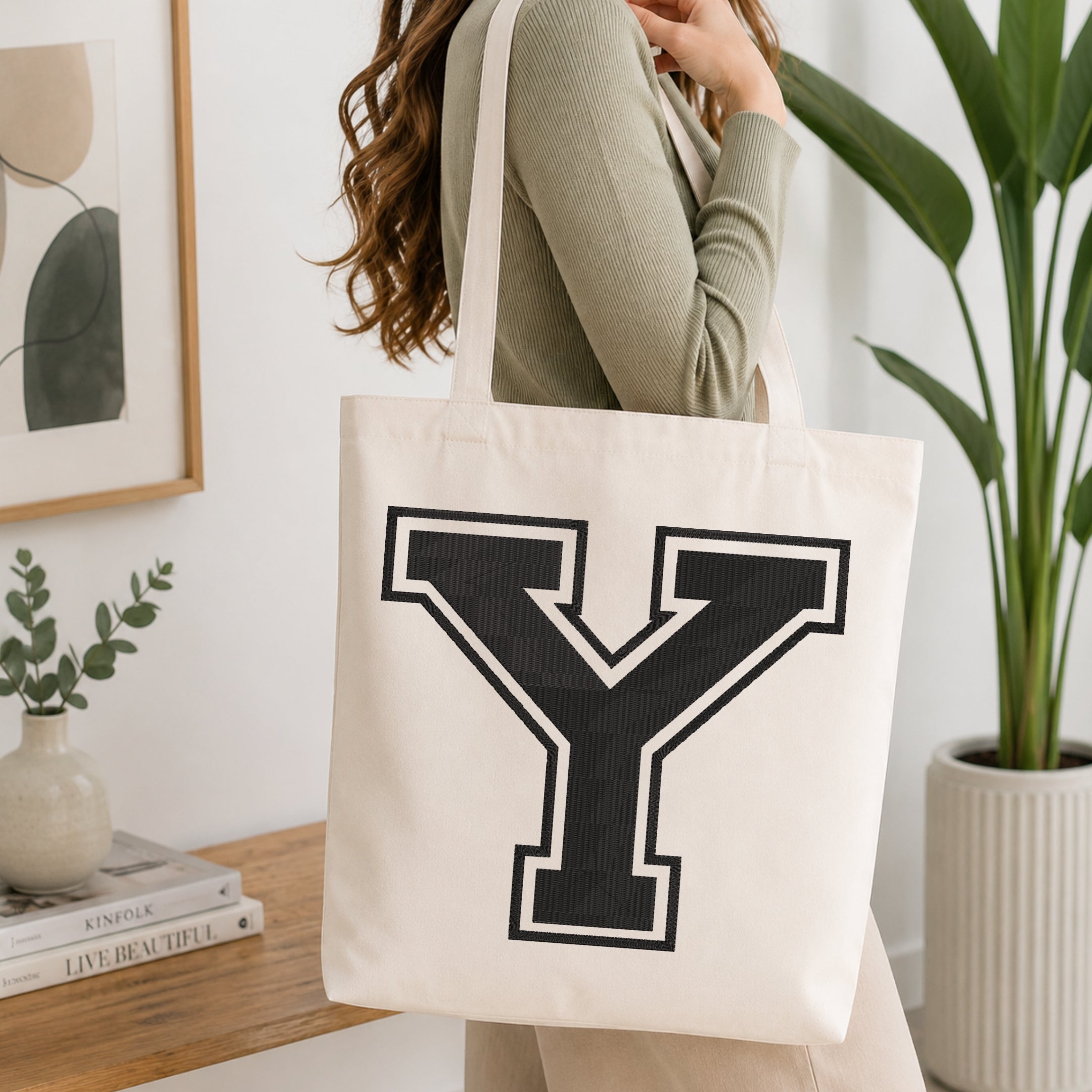 A woman wearing a sage green ribbed sweater carries a cream canvas tote bag with a large black collegiate letter 'Y' outlined in white on the front, set in a bright, modern room with a wooden console table, framed art, and a potted plant.