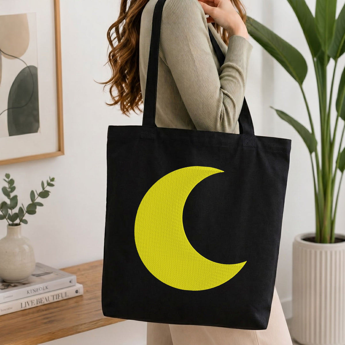 A person wearing a beige ribbed sweater carries a black canvas tote bag with a large bright yellow crescent moon graphic on the front, set in a bright, modern room with a wooden table, framed art, and a potted plant.