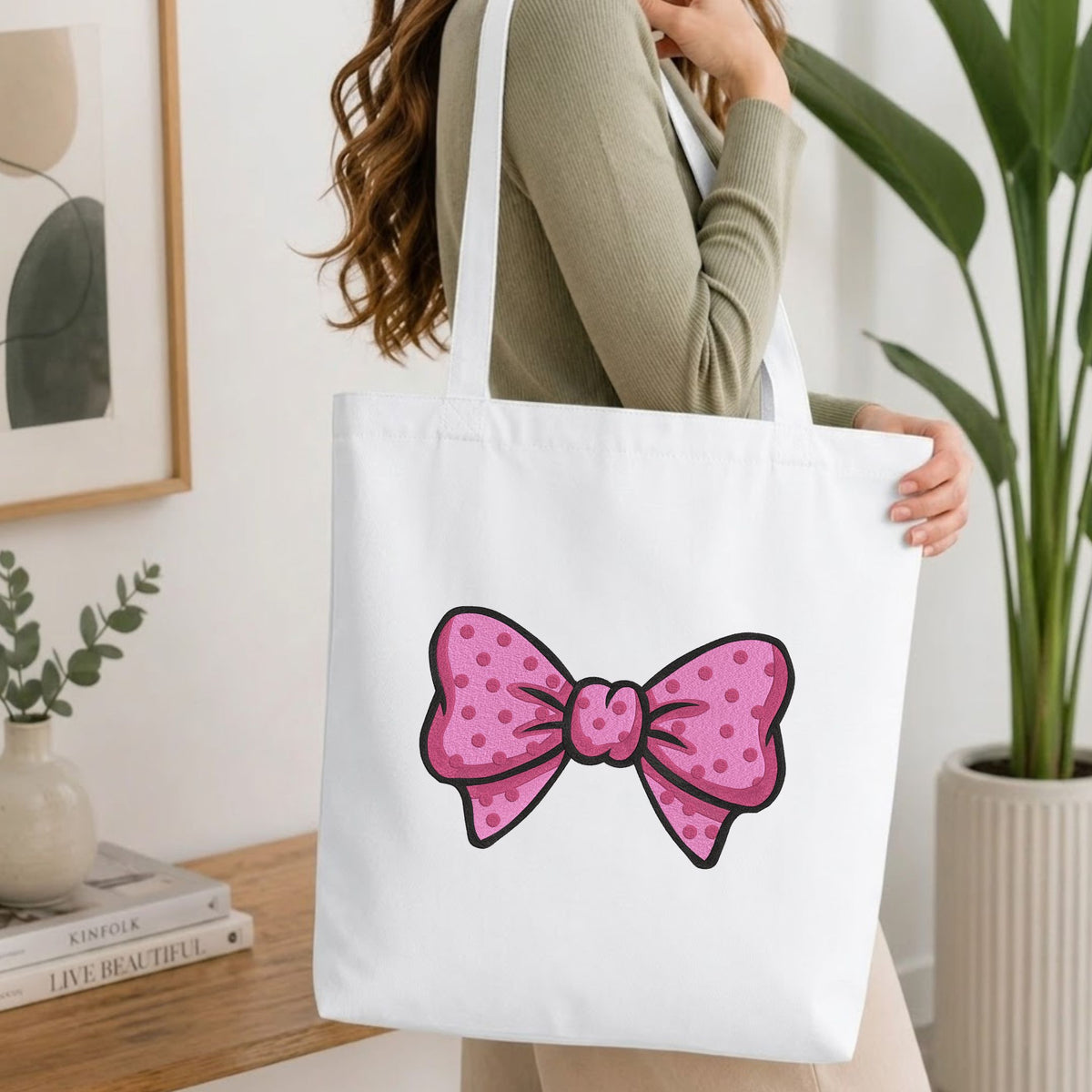 Woman carrying a white natural canvas tote bag featuring a pink polka dot bow machine embroidery design in a bright, modern interior setting   