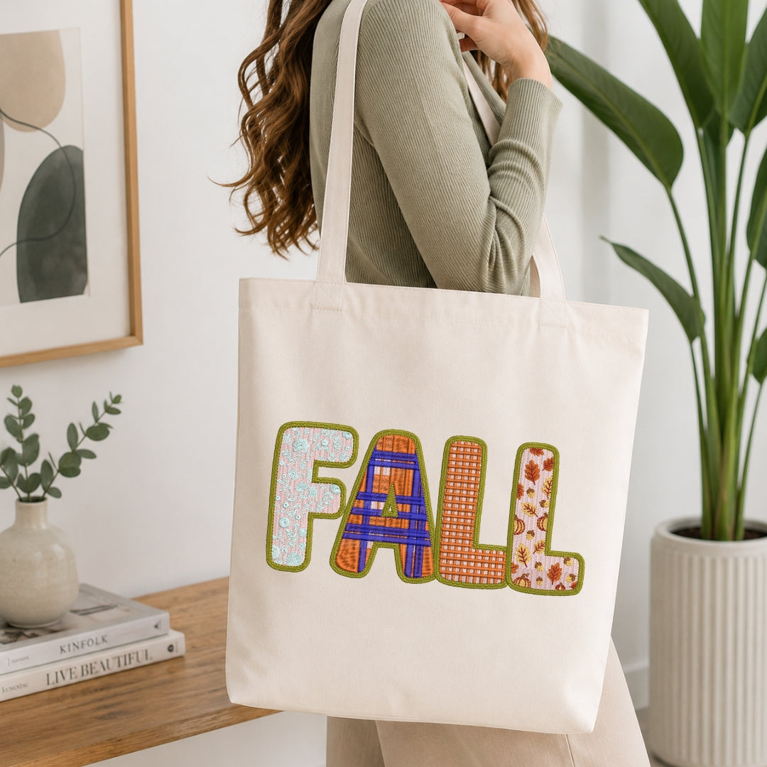 A woman wearing a sage green ribbed sweater carries a cream canvas tote bag with the word 'FALL' spelled out in colorful patterned letters. The scene is set in a bright, modern room with a wooden side table, a potted plant, and a framed print.