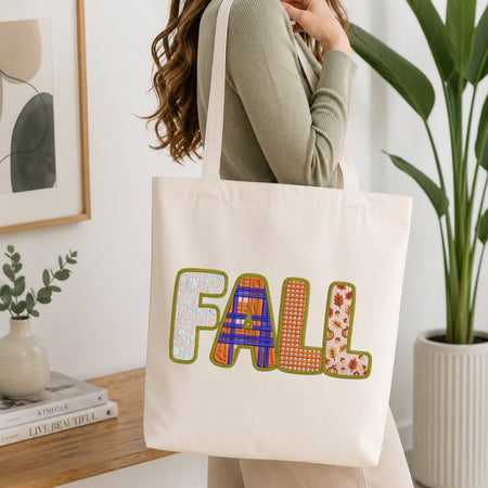 A woman wearing a sage green ribbed sweater carries a cream canvas tote bag with the word 'FALL' spelled out in colorful patterned letters. The scene is set in a bright, modern room with a wooden side table, a potted plant, and a framed print.
