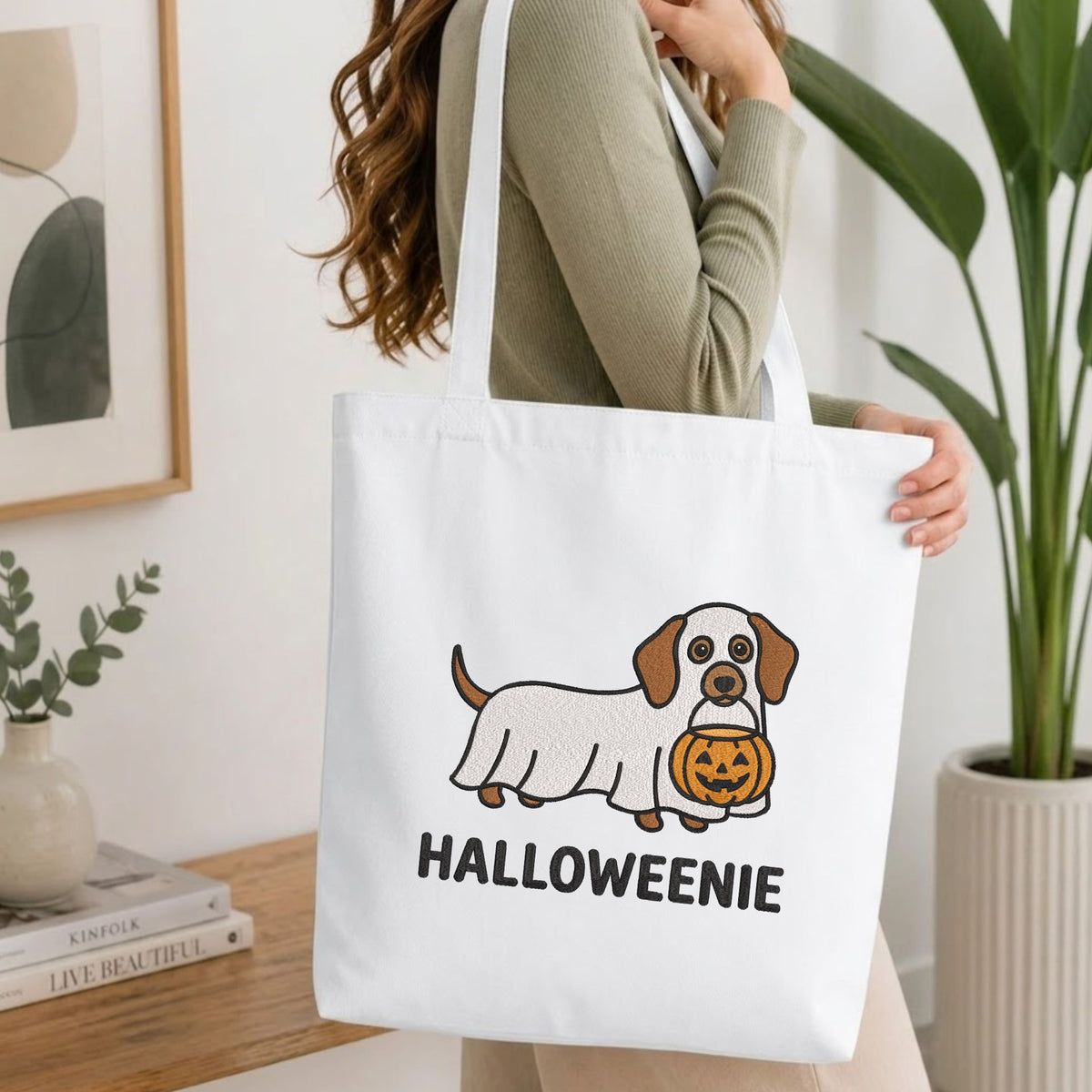 Folded white cotton t-shirt mockup featuring a "Halloweenie" dachshund ghost machine embroidery design, styled with cotton stems and a wooden cup