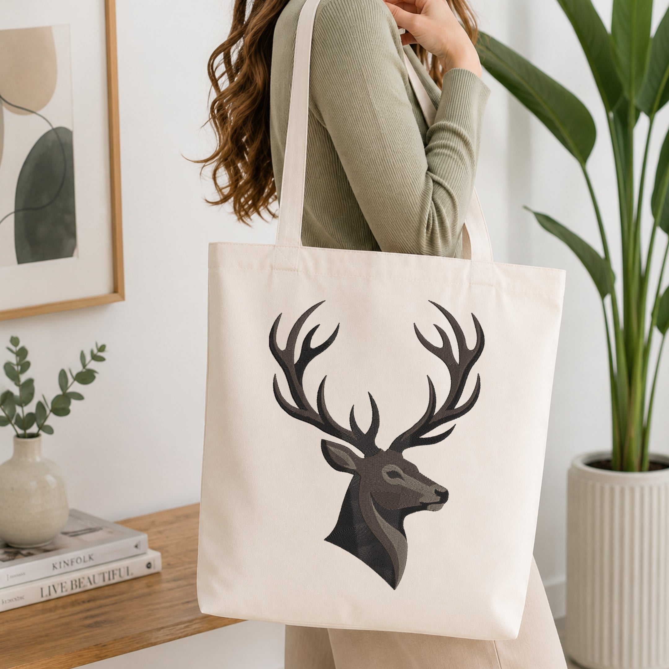 A woman wearing a sage green ribbed sweater carries a cream canvas tote bag with a large black and gray deer head graphic on the front, set in a bright, modern room with a wooden console table and a potted plant.